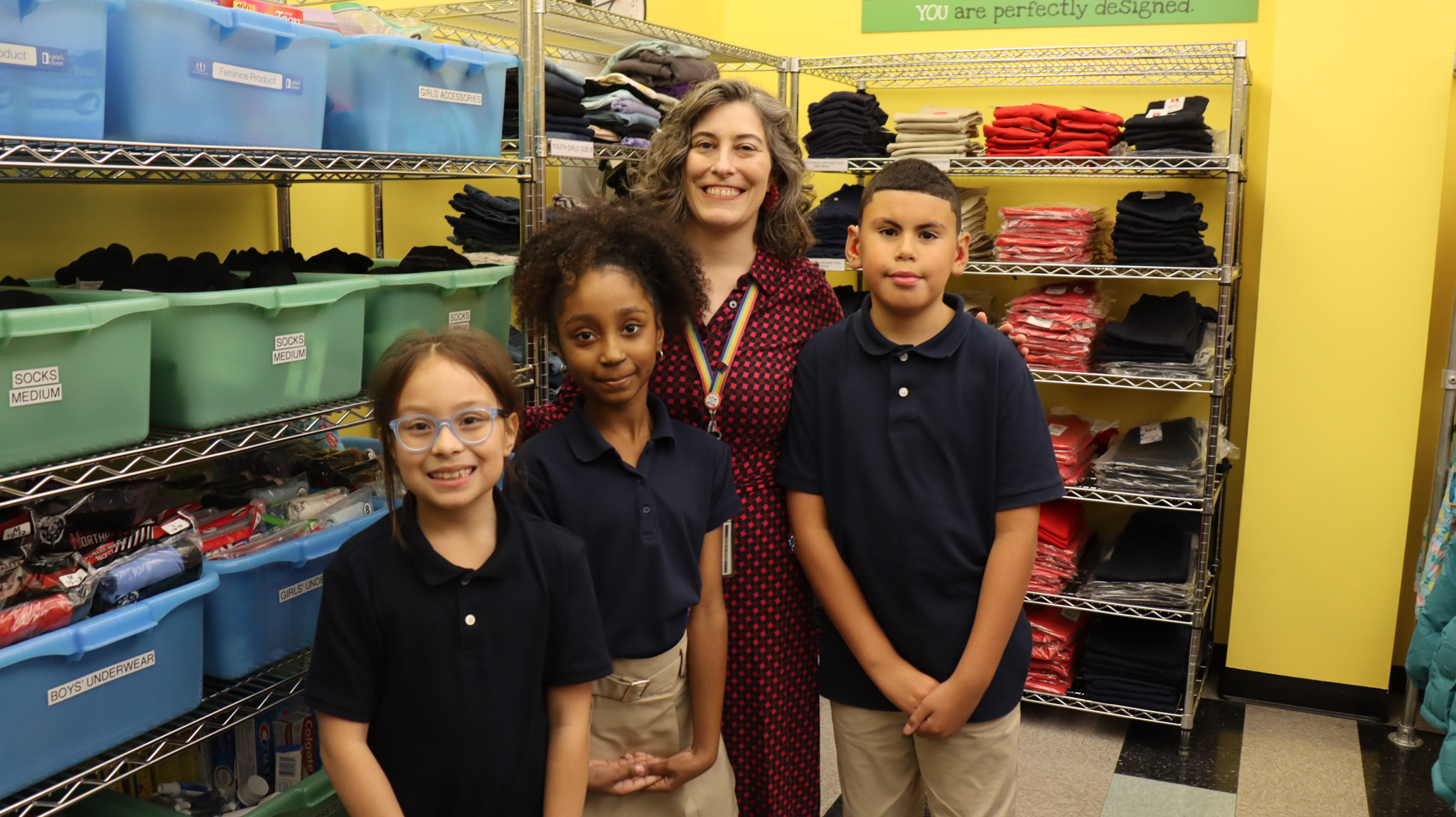 A group photo with three elementary school students and the school principal standing in a room filled with clothes and necessities for kids